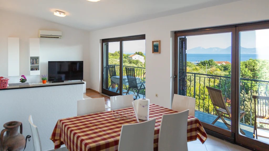 Open-plan living and dining area with a checked table, television and balcony doors onto a sea view
