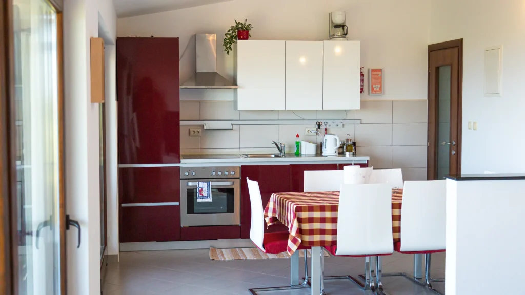 Fitted kitchen with dark red units, oven and sink, and a dining table with a red checked cloth and white chairs