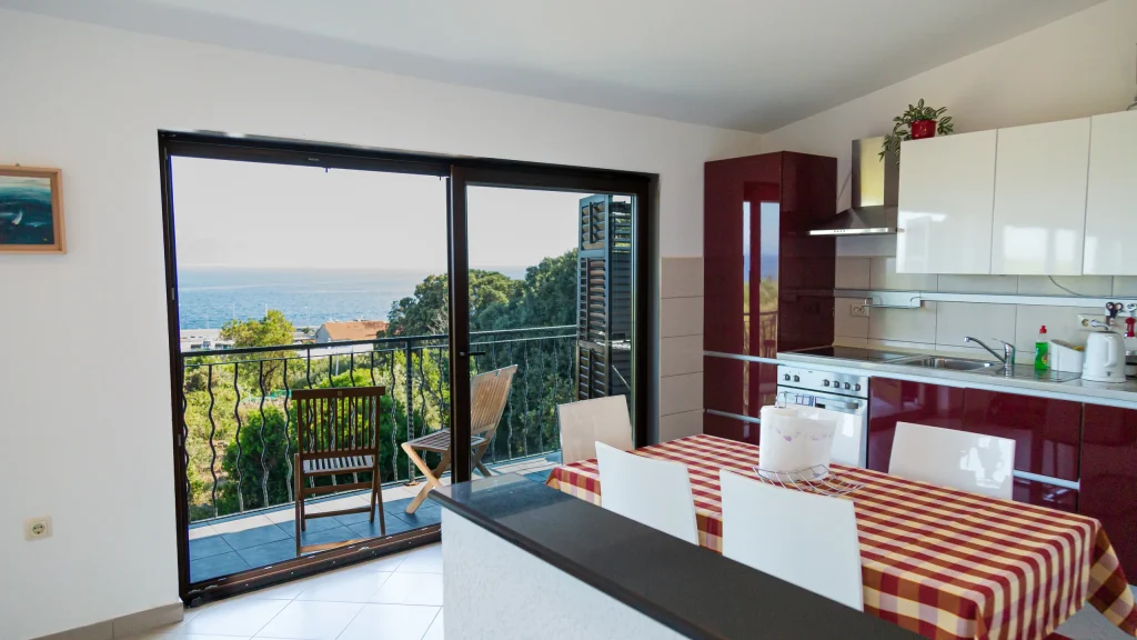 Kitchen with dark red units and a checked dining table beside sliding doors onto a balcony with a sea view