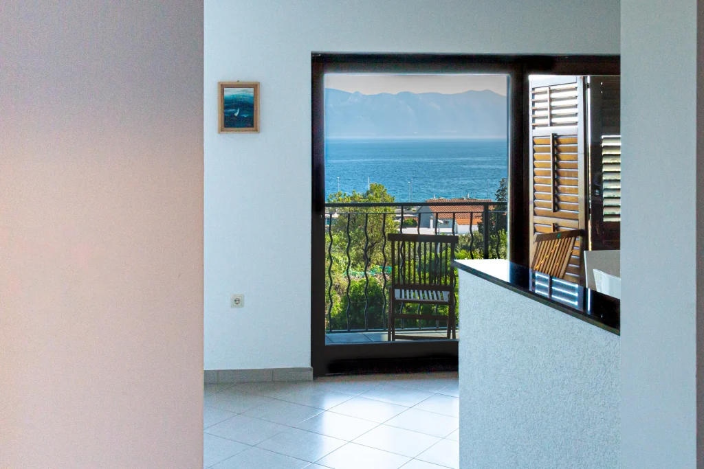 Room with a tiled floor, a framed seascape on the wall and a glass door onto a balcony, with sea and mountains beyond