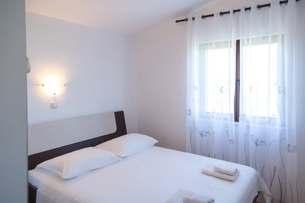 Double bed with white linen and folded towels, a wall light above the headboard and a curtained window