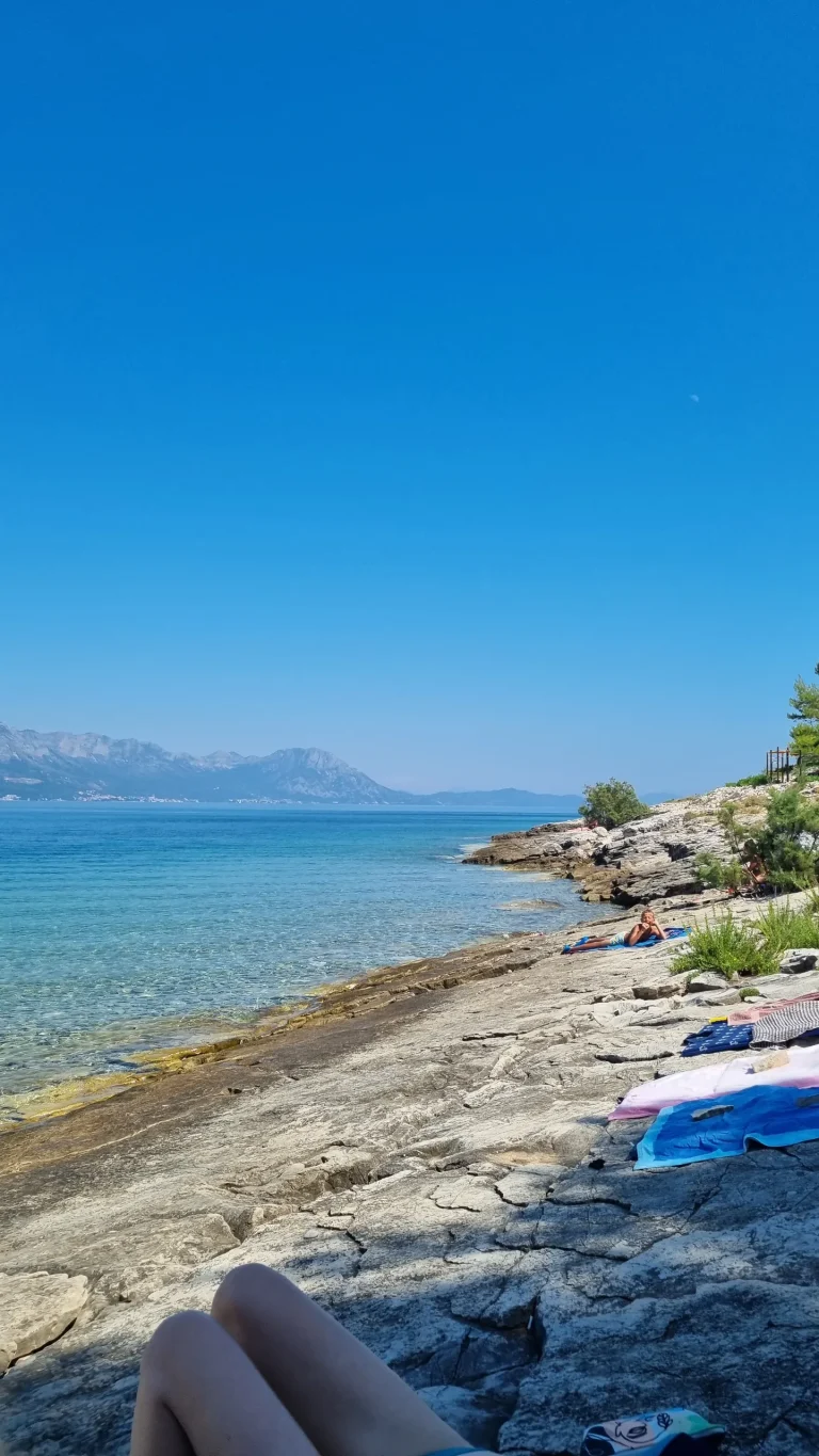 Flat rock shore with towels spread out beside clear turquoise water, pines to one side and mountains across the bay