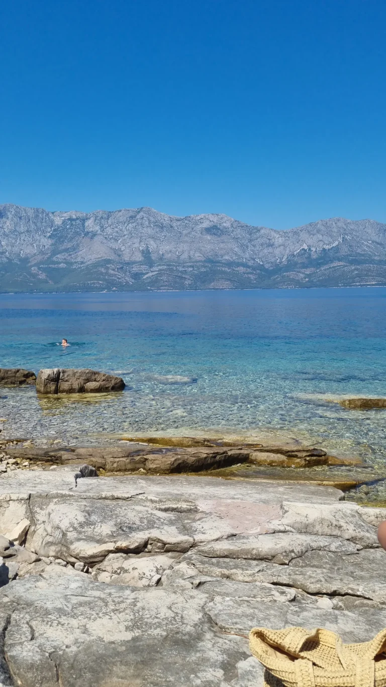 Rocky shoreline with clear turquoise water near Sućuraj, a swimmer offshore and a mountain ridge across the channel