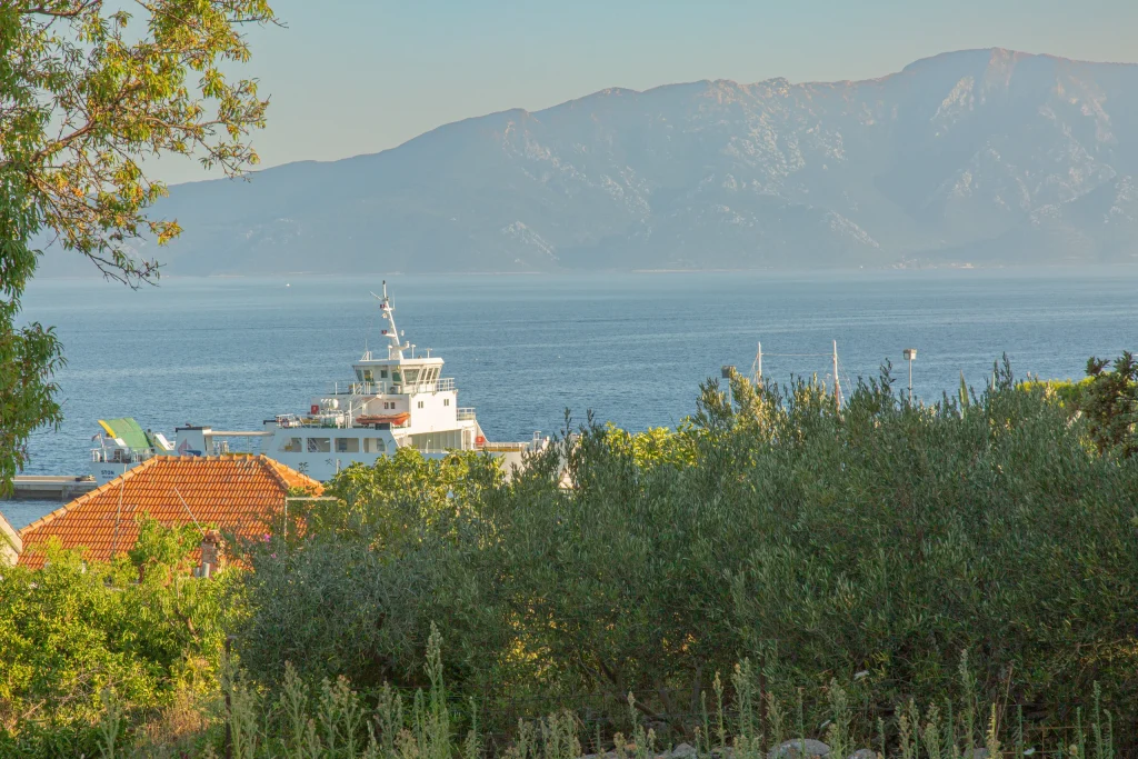 White car ferry moored at the quay below olive trees, with a hazy mountain ridge across the channel