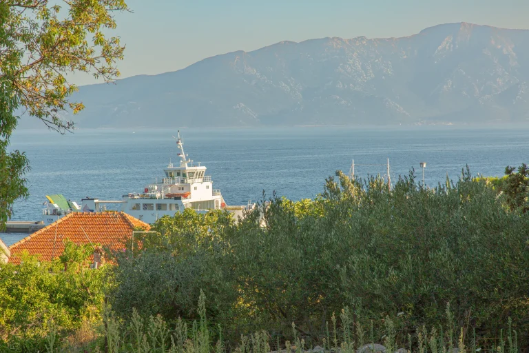 White car ferry moored at the quay below olive trees, with a hazy mountain ridge across the channel