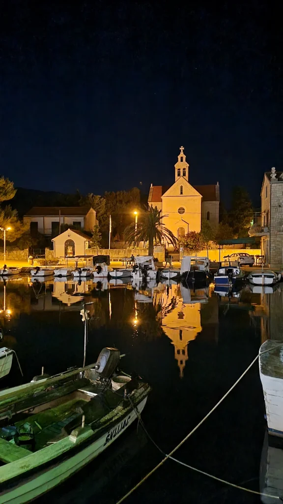 Lit church and stone houses above a small harbour at night, moored boats reflected in still water