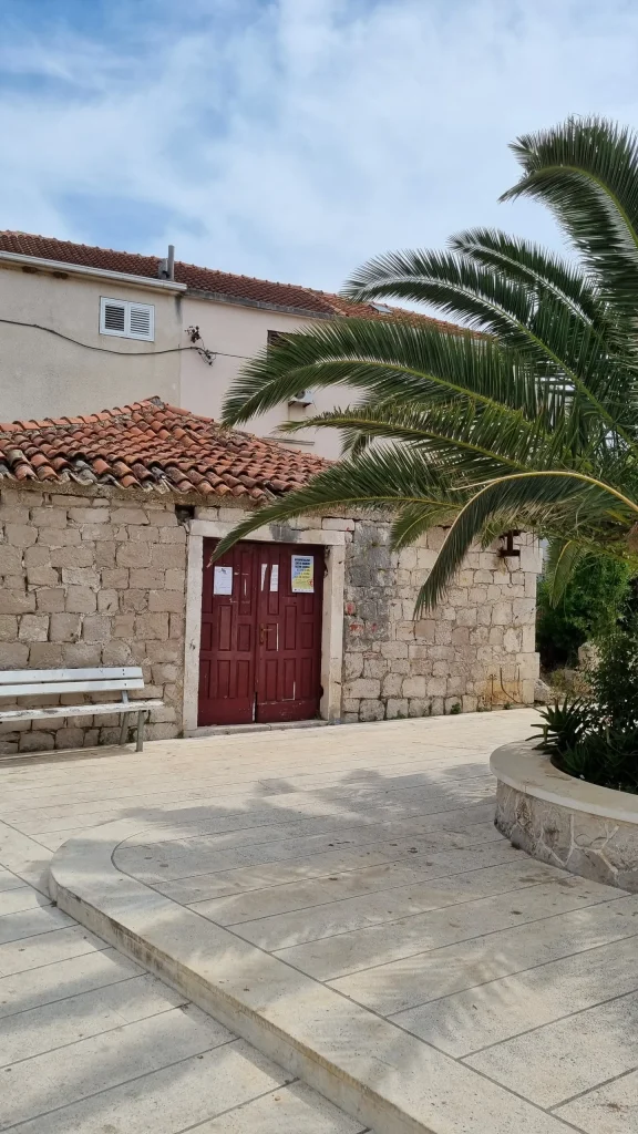 Paved square in Sućuraj with a stone building, red wooden doors, a white bench and a palm in a stone planter