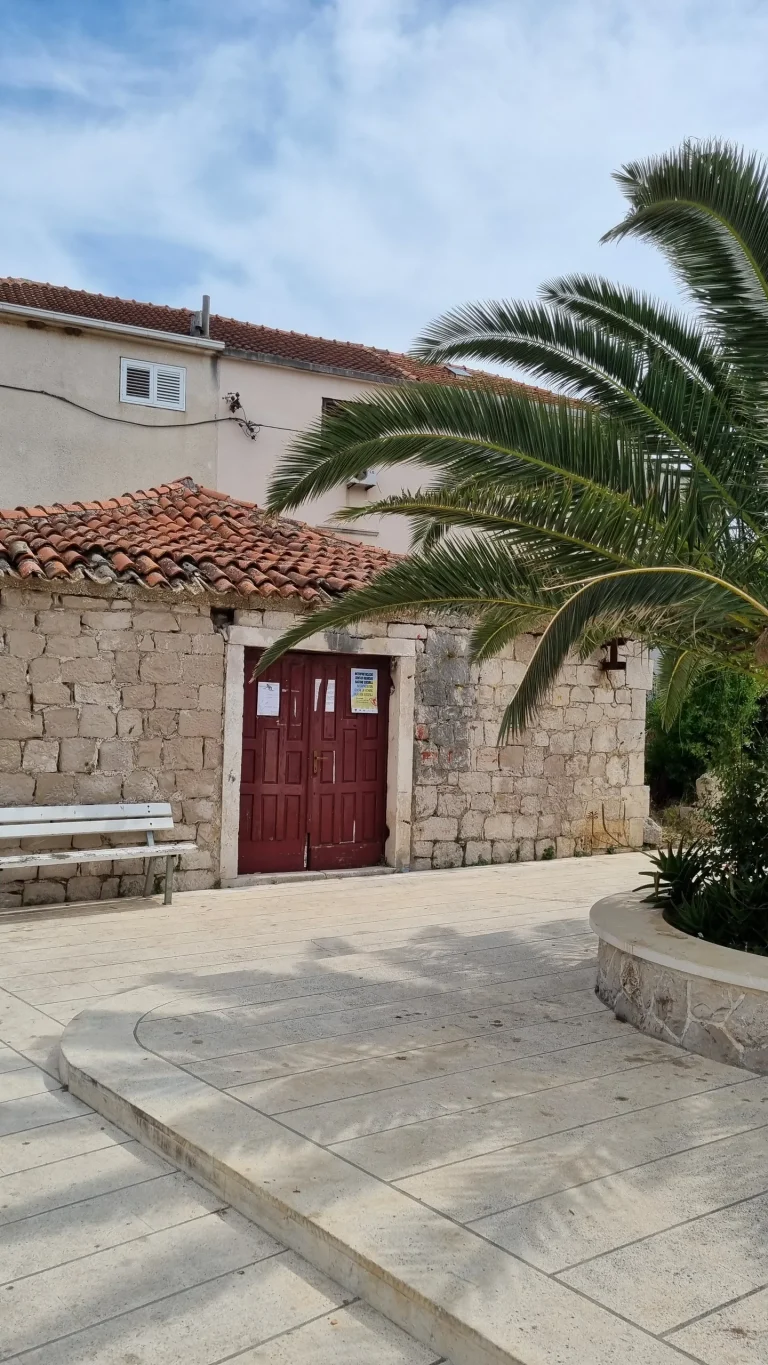Paved square in Sućuraj with a stone building, red wooden doors, a white bench and a palm in a stone planter