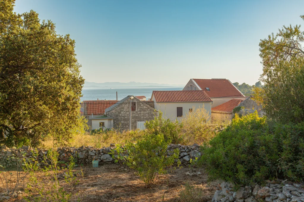 Village houses with terracotta roofs seen from a garden with dry stone walls, the sea and distant mainland beyond