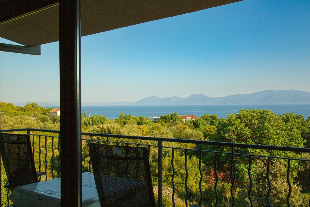 Table and two chairs on a shaded balcony behind a metal railing, looking over greenery to the sea and mountains