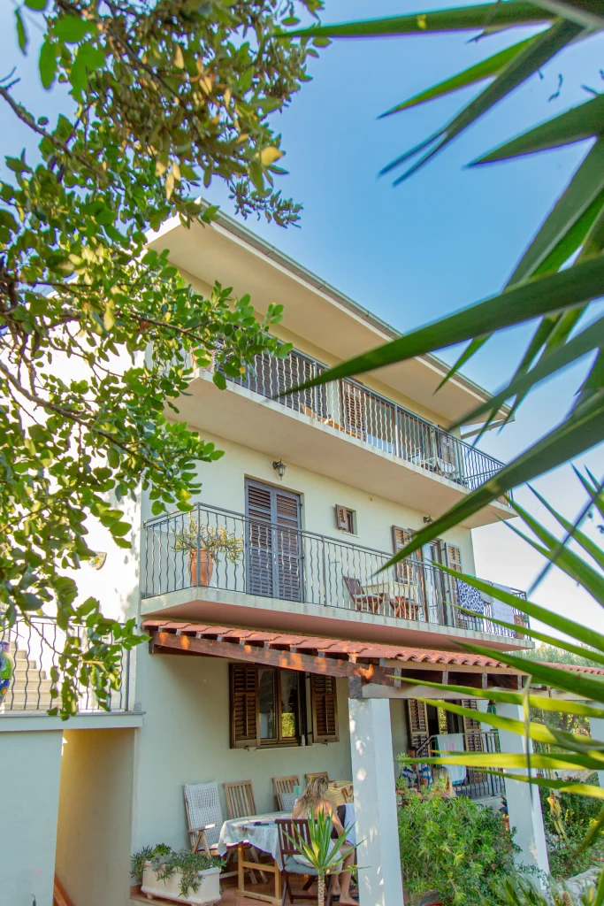 House seen from the garden through leafy branches, balconies with shutters above a terrace where guests are sitting