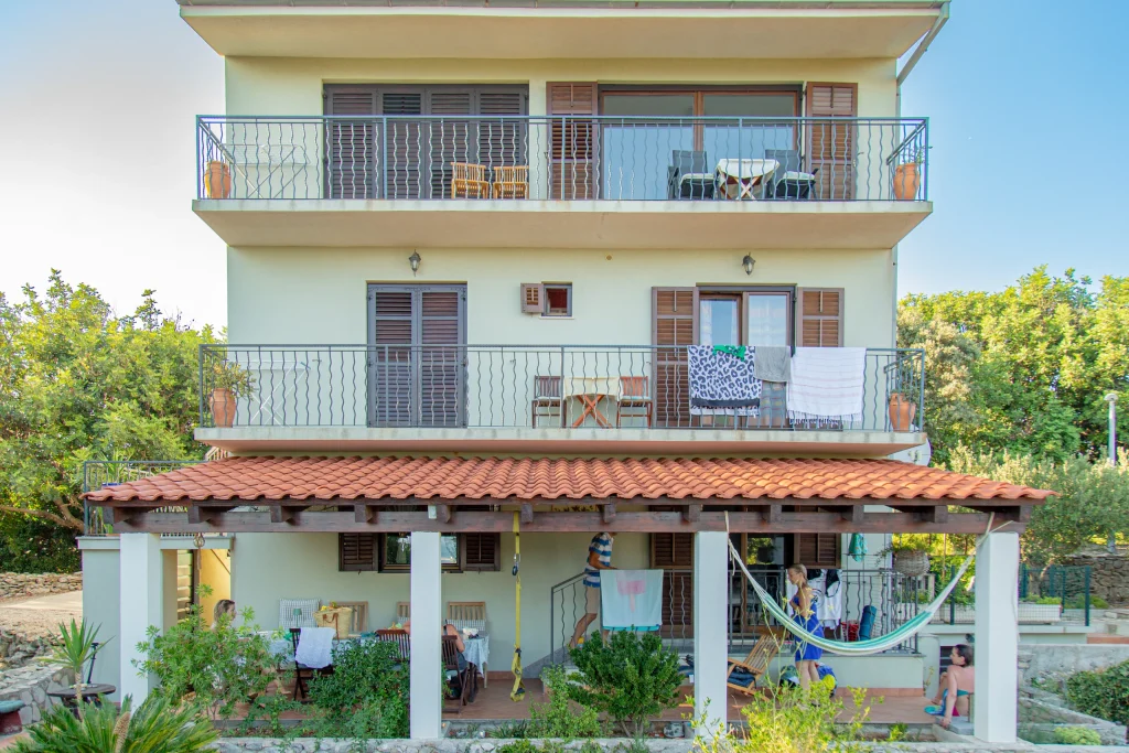 Three-storey house with two balconies above a tiled-roof terrace where guests sit at a table, with a hammock alongside